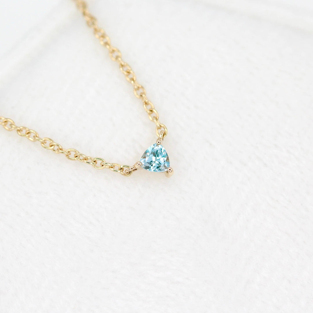 The March Tiny Trillion Birthstone Necklace with Aqua Spinel in Yellow Gold atop of a white velvet ring box