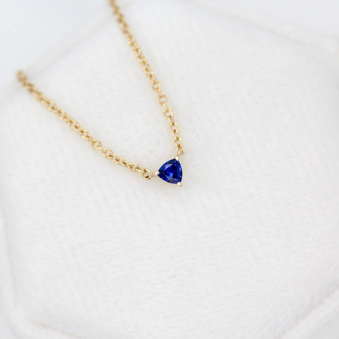 The September Birthstone Tiny Trillion Necklace with Blue Sapphire in Yellow Gold against a white velvet ring box