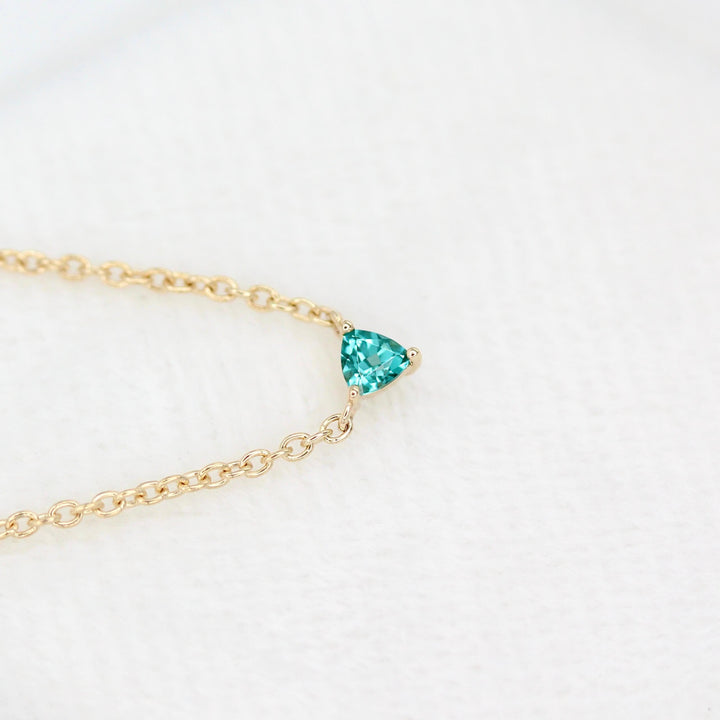 The December Birthstone Tiny Trillion Necklace with Paraiba Spinel in Yellow Gold against a white velvet ring box