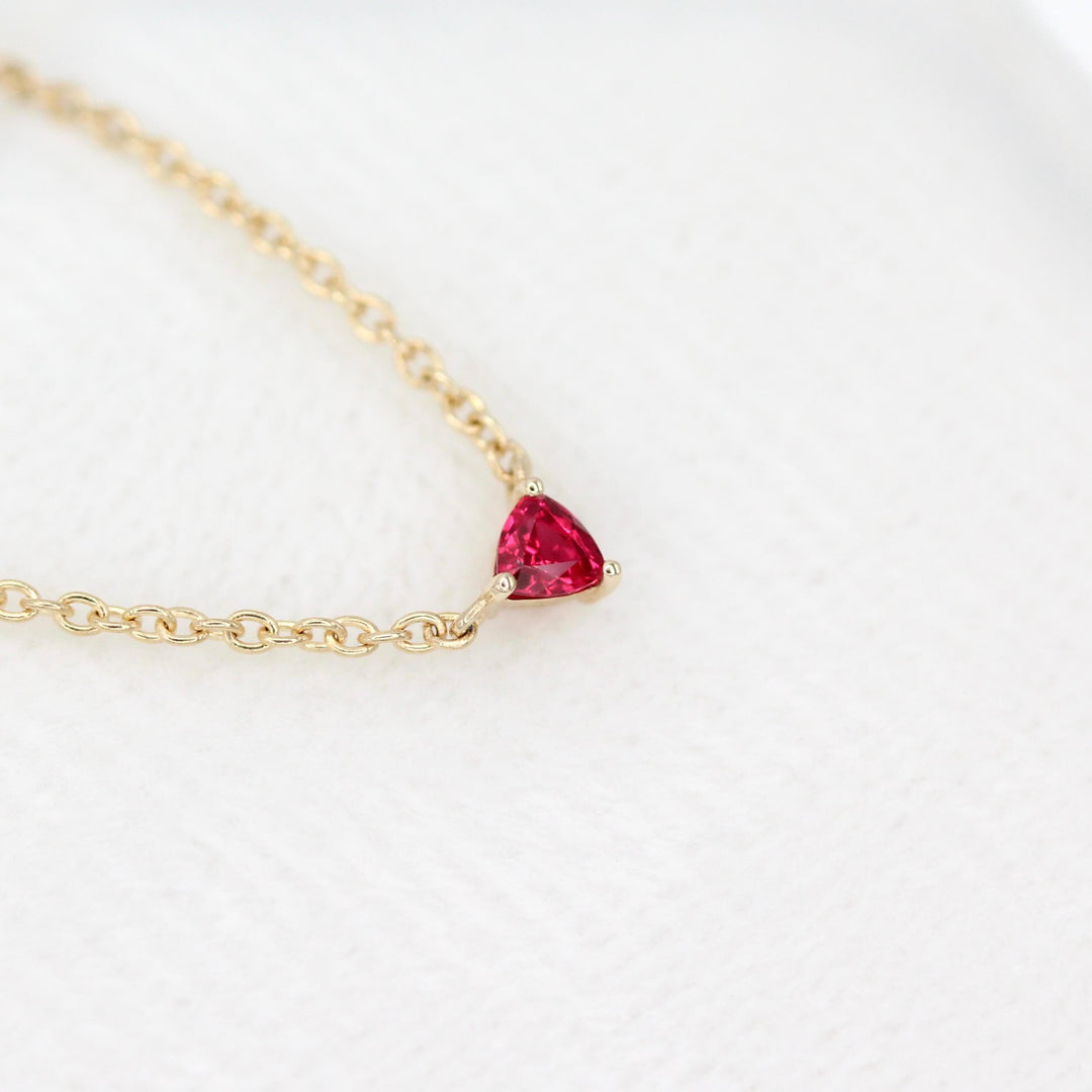 The July Birthstone Tiny Trillion Necklace with Ruby in Yellow Gold against a white velvet ring box